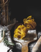 Two nests of uncooked tagliatelle pasta are placed on a wooden cutting board dusted with flour. Rosemary sprigs are tied together beside the board on a linen cloth. A rolling pin is partially visible on the right, while a jar holding bundled utensils can be seen in the background.