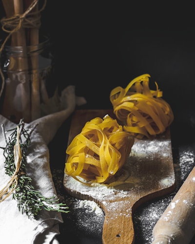 Two nests of uncooked tagliatelle pasta are placed on a wooden cutting board dusted with flour. Rosemary sprigs are tied together beside the board on a linen cloth. A rolling pin is partially visible on the right, while a jar holding bundled utensils can be seen in the background.