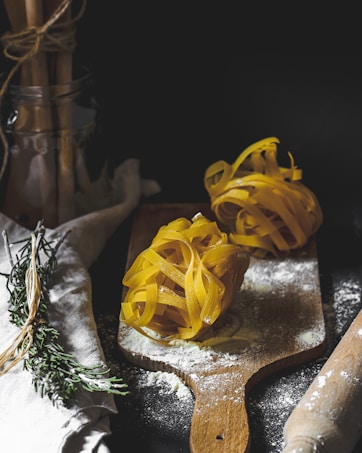 Two nests of uncooked tagliatelle pasta are placed on a wooden cutting board dusted with flour. Rosemary sprigs are tied together beside the board on a linen cloth. A rolling pin is partially visible on the right, while a jar holding bundled utensils can be seen in the background.