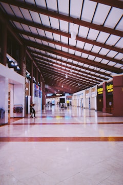 A spacious, well-lit indoor corridor with a high, wooden beam ceiling. The floor features a polished, light-colored tile with dark red stripes. Various stores and signs line the sides, each displaying bright, colorful signage. A few people are scattered in the distance, walking or standing, and there is a janitor sweeping on the left.