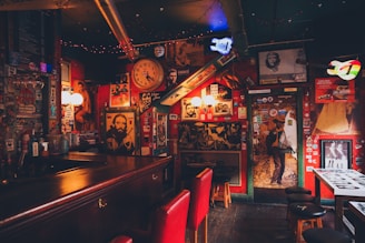 An eclectic bar interior filled with vintage posters, photographs, and memorabilia covering the walls. Dim lighting creates a cozy, intimate atmosphere. A large wall clock, colorful lights, and red cushioned bar stools contribute to the vibrant yet nostalgic ambiance.