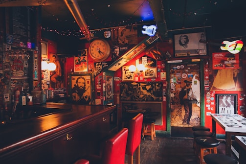 An eclectic bar interior filled with vintage posters, photographs, and memorabilia covering the walls. Dim lighting creates a cozy, intimate atmosphere. A large wall clock, colorful lights, and red cushioned bar stools contribute to the vibrant yet nostalgic ambiance.