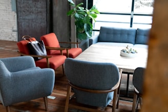 A modern living room setup includes comfortable armchairs in blue and red, a large wooden table in the center, and a gray sofa against a backdrop of large windows. A black leather handbag rests on a red chair, while a potted plant is seen in the corner. The space is warmly lit, with wooden floors and a decorative bowl on the table.
