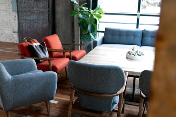 A modern living room setup includes comfortable armchairs in blue and red, a large wooden table in the center, and a gray sofa against a backdrop of large windows. A black leather handbag rests on a red chair, while a potted plant is seen in the corner. The space is warmly lit, with wooden floors and a decorative bowl on the table.