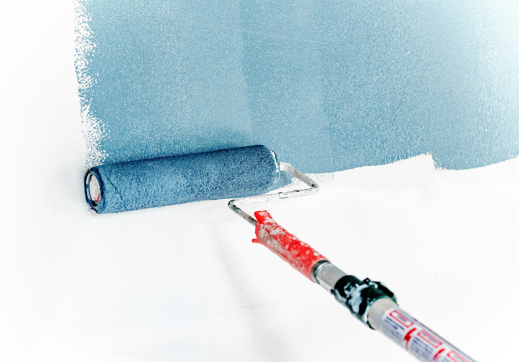 Blue paint being rolled onto an empty white wall with an extended paint roller