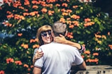 A couple embraces in front of a lush backdrop filled with vibrant orange flowers. The woman, wearing sunglasses, looks directly at the camera with a joyful smile while her hand rests affectionately around the man's neck. The man, seen from the back, wears a white shirt with striped sleeves. The setting appears warm and inviting with greenery surrounding the couple.