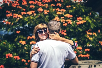 A couple embraces in front of a lush backdrop filled with vibrant orange flowers. The woman, wearing sunglasses, looks directly at the camera with a joyful smile while her hand rests affectionately around the man's neck. The man, seen from the back, wears a white shirt with striped sleeves. The setting appears warm and inviting with greenery surrounding the couple.