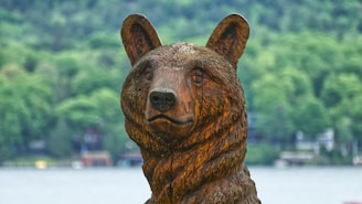 A large, intricately carved wooden bear statue with detailed facial features stands in an outdoor setting. The background consists of a blurred view of lush green trees and a body of water, suggesting a natural environment.