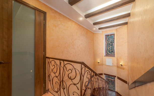 A staircase with ornate metal railings and wooden handrail, leading to an upper floor. The walls are painted in warm, peach tones with texture, and the ceiling features exposed dark wooden beams. There are two wall sconces providing warm lighting, and a window with decorative ironwork.