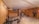 A well-lit home gym with wooden floors and beige walls is equipped with various exercise machines, including a bench press, stationary bike, and multi-gym station. A beige sofa is placed against one wall near an air conditioning unit and a small window.