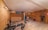 A well-lit home gym with wooden floors and beige walls is equipped with various exercise machines, including a bench press, stationary bike, and multi-gym station. A beige sofa is placed against one wall near an air conditioning unit and a small window.