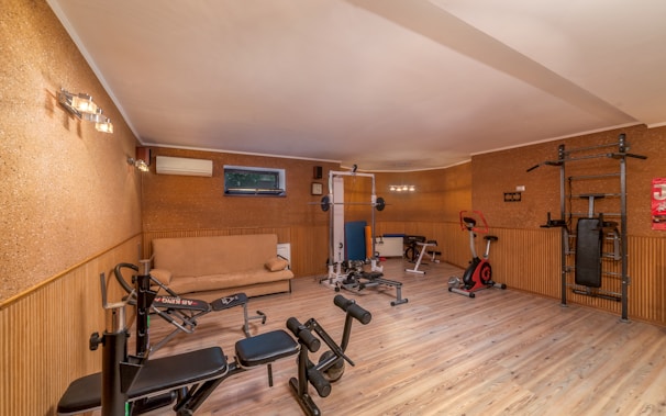 A well-lit home gym with wooden floors and beige walls is equipped with various exercise machines, including a bench press, stationary bike, and multi-gym station. A beige sofa is placed against one wall near an air conditioning unit and a small window.