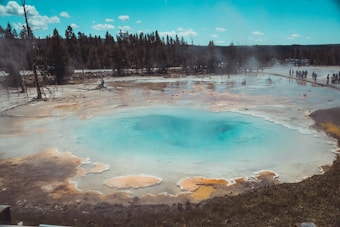 A vibrant geothermal hot spring with a striking turquoise center and surrounded by multicolored mineral deposits. Tall trees and a clear blue sky enhance the natural scenery, while steam rises from the spring, creating a mystical atmosphere. Visitors can be seen at a distance, observing the spectacle from a pathway.