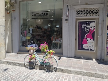 A shopfront displays a sign for 'Catwalk Accessories' with large windows showing various bags and items inside. In front of the shop, a vintage bicycle is adorned with baskets filled with vibrant flowers, adding a charming touch to the scene. Next to the shop entrance is a poster featuring an illustration of a fashionable woman holding a pink handbag.