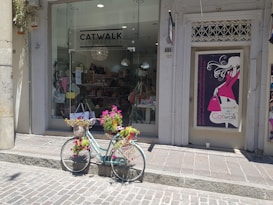 A shopfront displays a sign for 'Catwalk Accessories' with large windows showing various bags and items inside. In front of the shop, a vintage bicycle is adorned with baskets filled with vibrant flowers, adding a charming touch to the scene. Next to the shop entrance is a poster featuring an illustration of a fashionable woman holding a pink handbag.