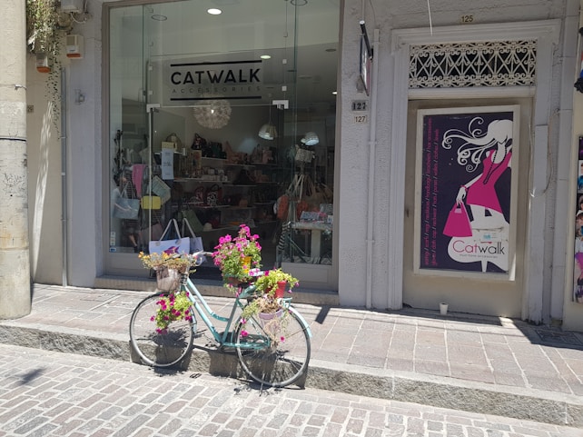 A shopfront displays a sign for 'Catwalk Accessories' with large windows showing various bags and items inside. In front of the shop, a vintage bicycle is adorned with baskets filled with vibrant flowers, adding a charming touch to the scene. Next to the shop entrance is a poster featuring an illustration of a fashionable woman holding a pink handbag.
