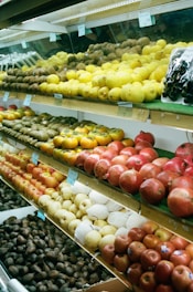 A variety of fruits are displayed on multiple shelves inside a grocery store. The shelves are neatly arranged with lemons, kiwis, pomegranates, apples, pears, and avocados. The presentation is orderly, with each fruit type organized in distinct sections. Clear price tags are visible above some of the fruits, indicating a commercial setting.