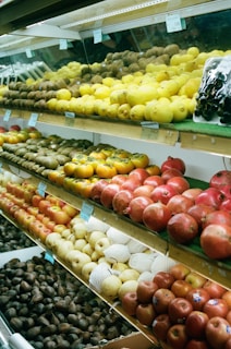 A variety of fruits are displayed on multiple shelves inside a grocery store. The shelves are neatly arranged with lemons, kiwis, pomegranates, apples, pears, and avocados. The presentation is orderly, with each fruit type organized in distinct sections. Clear price tags are visible above some of the fruits, indicating a commercial setting.