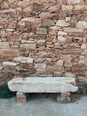 A rustic stone bench set against a textured stone wall composed of variously sized rocks and bricks. The bench appears weathered with a light hue, contrasting with the darker background. A patch of green foliage peeks out from the side, adding a touch of nature to the otherwise earthy scene.