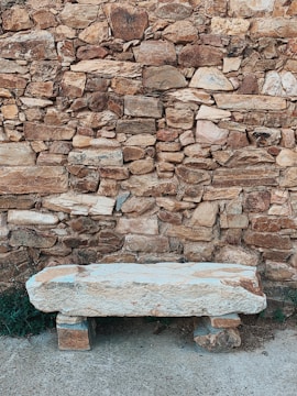 A rustic stone bench set against a textured stone wall composed of variously sized rocks and bricks. The bench appears weathered with a light hue, contrasting with the darker background. A patch of green foliage peeks out from the side, adding a touch of nature to the otherwise earthy scene.