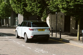 A white Range Rover is parked on a cobblestone street lined with tall green trees. The vehicle's rear license plate is visible, and shadows from the trees are cast on the road. The scene is set in an urban area with bollards and buildings visible in the background.