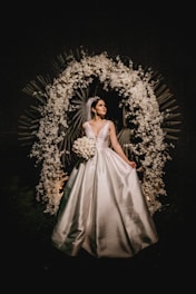 A bride in a white gown stands elegantly under a decorative arch made of white flowers and leaves. The setting is dark, making the bride and floral arrangement the focal points.