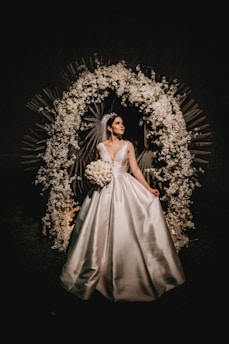 A bride in a white gown stands elegantly under a decorative arch made of white flowers and leaves. The setting is dark, making the bride and floral arrangement the focal points.