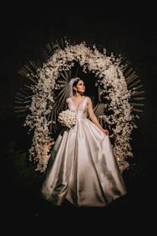 A bride in a white gown stands elegantly under a decorative arch made of white flowers and leaves. The setting is dark, making the bride and floral arrangement the focal points.