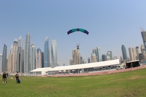 A tandem skydiver is descending with a colorful parachute above a grassy area in an urban environment. In the background, a skyline of modern skyscrapers stands tall under a clear blue sky. A white building and a few people on the ground can also be seen.