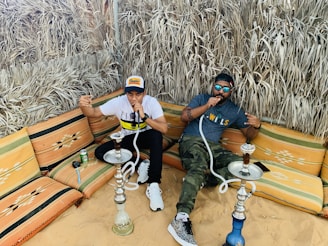 Two people are sitting on colorful cushioned seating in a sandy outdoor area, enjoying smoking shisha. They are surrounded by dried grass walls, creating a rustic atmosphere. Each person is holding a shisha hose, and there is a can and some items placed on the seating next to them. Both individuals are dressed casually with one in a cap and the other wearing sunglasses.