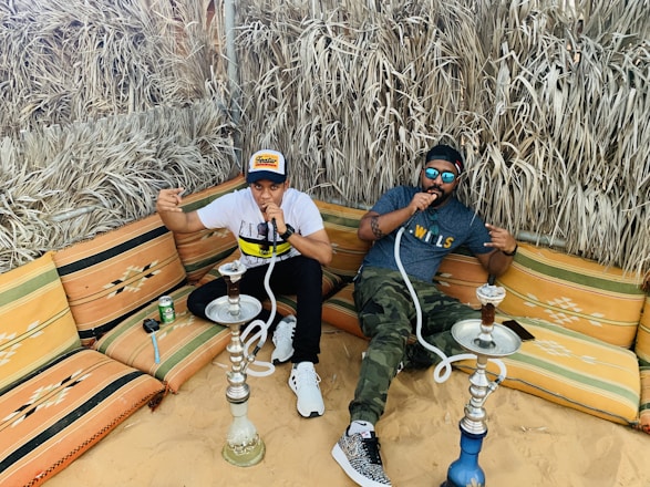 Two people are sitting on colorful cushioned seating in a sandy outdoor area, enjoying smoking shisha. They are surrounded by dried grass walls, creating a rustic atmosphere. Each person is holding a shisha hose, and there is a can and some items placed on the seating next to them. Both individuals are dressed casually with one in a cap and the other wearing sunglasses.