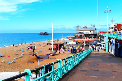 A vibrant beachfront scene showing a sandy area with numerous deck chairs scattered across the beach. People are walking and relaxing while the sea stretches into the horizon. To the right, there is a bustling promenade with various shops and cafes. An old pier structure is visible in the background, and buildings rise along the coastal edge.