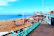 A vibrant beachfront scene showing a sandy area with numerous deck chairs scattered across the beach. People are walking and relaxing while the sea stretches into the horizon. To the right, there is a bustling promenade with various shops and cafes. An old pier structure is visible in the background, and buildings rise along the coastal edge.
