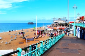 A vibrant beachfront scene showing a sandy area with numerous deck chairs scattered across the beach. People are walking and relaxing while the sea stretches into the horizon. To the right, there is a bustling promenade with various shops and cafes. An old pier structure is visible in the background, and buildings rise along the coastal edge.