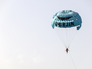 A person is parasailing with a large blue parachute that features a smiling face and the word 'ALMARJAN' printed on it, set against a clear sky.
