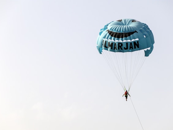A person is parasailing with a large blue parachute that features a smiling face and the word 'ALMARJAN' printed on it, set against a clear sky.