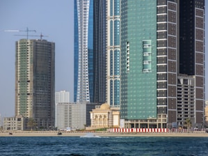 A waterfront cityscape with modern high-rise buildings, including a green building with balconies and a construction site with cranes. In the foreground, there are rippling water waves, and a sandy beach is visible at the base of the buildings.