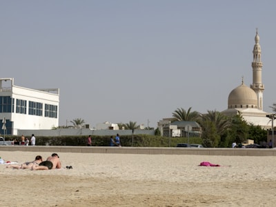 A sandy beach with a pair of people lying on towels at the forefront, sunbathing. In the background, there is a mosque with a prominent dome and minaret surrounded by greenery. The sky is clear, and other people are walking and relaxing nearby.