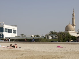 A sandy beach with a pair of people lying on towels at the forefront, sunbathing. In the background, there is a mosque with a prominent dome and minaret surrounded by greenery. The sky is clear, and other people are walking and relaxing nearby.