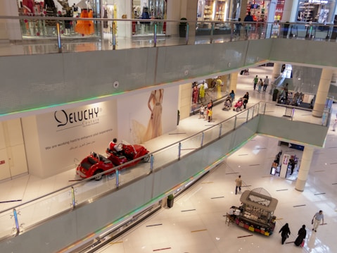 A multi-level shopping mall with several people walking around. There are clothing stores with glass fronts, a bright display of mannequins, and a few people pushing strollers. A red vehicle resembling a decorative ride is placed along a walkway. An advertisement for a fashion brand is visible on the wall.