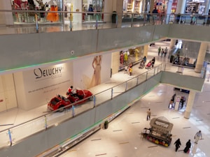 A multi-level shopping mall with several people walking around. There are clothing stores with glass fronts, a bright display of mannequins, and a few people pushing strollers. A red vehicle resembling a decorative ride is placed along a walkway. An advertisement for a fashion brand is visible on the wall.