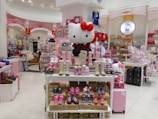 A colorful retail store filled with an assortment of Hello Kitty merchandise, including shoes, clothing, bags, and plush toys. The display in the center shows various styles of kids' footwear, with a large plush toy of Hello Kitty wearing a red bow sitting prominently on top. The store has a bright and cheerful atmosphere with pink and white as the dominant colors. Numerous Hello Kitty themed items are arranged neatly on shelves and tables throughout the store.