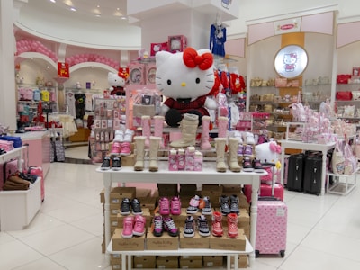 A colorful retail store filled with an assortment of Hello Kitty merchandise, including shoes, clothing, bags, and plush toys. The display in the center shows various styles of kids' footwear, with a large plush toy of Hello Kitty wearing a red bow sitting prominently on top. The store has a bright and cheerful atmosphere with pink and white as the dominant colors. Numerous Hello Kitty themed items are arranged neatly on shelves and tables throughout the store.