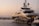 A luxurious yacht is docked at a marina, surrounded by calm waters and smaller boats visible in the background. The sky is tinged with warm hues of a sunset, creating a serene atmosphere.