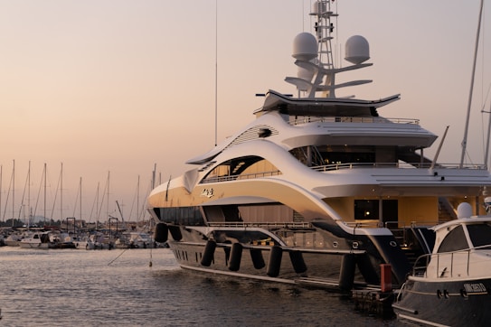 A luxurious yacht is docked at a marina, surrounded by calm waters and smaller boats visible in the background. The sky is tinged with warm hues of a sunset, creating a serene atmosphere.