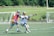 Two lacrosse players are actively engaging in a game on a grassy field, one wearing a colorful uniform and the other in white and blue, both equipped with helmets and sticks. There are goals and a lush green background indicating an outdoor sports environment.