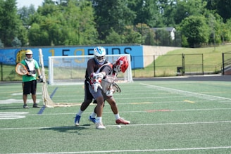 Two lacrosse players engage in a match on a synthetic field. One player attempts to take control of the ball while the other defends, both wearing helmets and protective gear. A goalie stands in front of the goal in the background, prepared to block any incoming shots. Trees and a fence provide a backdrop to the sports scene.