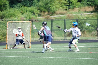 A group of young men are playing a game of lacrosse on an outdoor field. One player, dressed in a white and blue jersey, is running towards the goal while holding a lacrosse stick. The goalie, standing in front of the net, is wearing protective gear and is ready to defend. Another player in a brightly colored helmet is closely following the player with the ball, also holding a lacrosse stick. The field is green and surrounded by a fence with trees in the background.