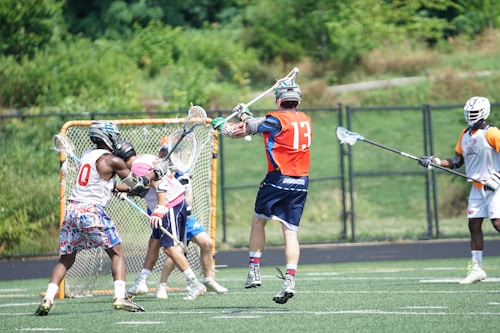 A group of young athletes engaged in a lacrosse game on a field. One player in a red jersey jumps with a lacrosse stick raised high, while others in various colored jerseys, including white and orange, actively participate close to a goal net. The background shows green bushes and a fence, indicating an outdoor environment.