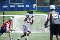Several athletes wearing protective gear are actively playing lacrosse on a field. A scoreboard is visible in the background, suggesting an organized game setting. The players are dressed in colorful uniforms, and their expressions reflect focus and determination.